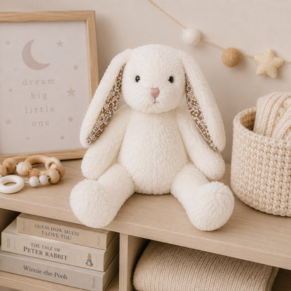 Eclectic Bun-Bun white plush bunny toy with floral ear lining sitting on a wooden nursery shelf with books.