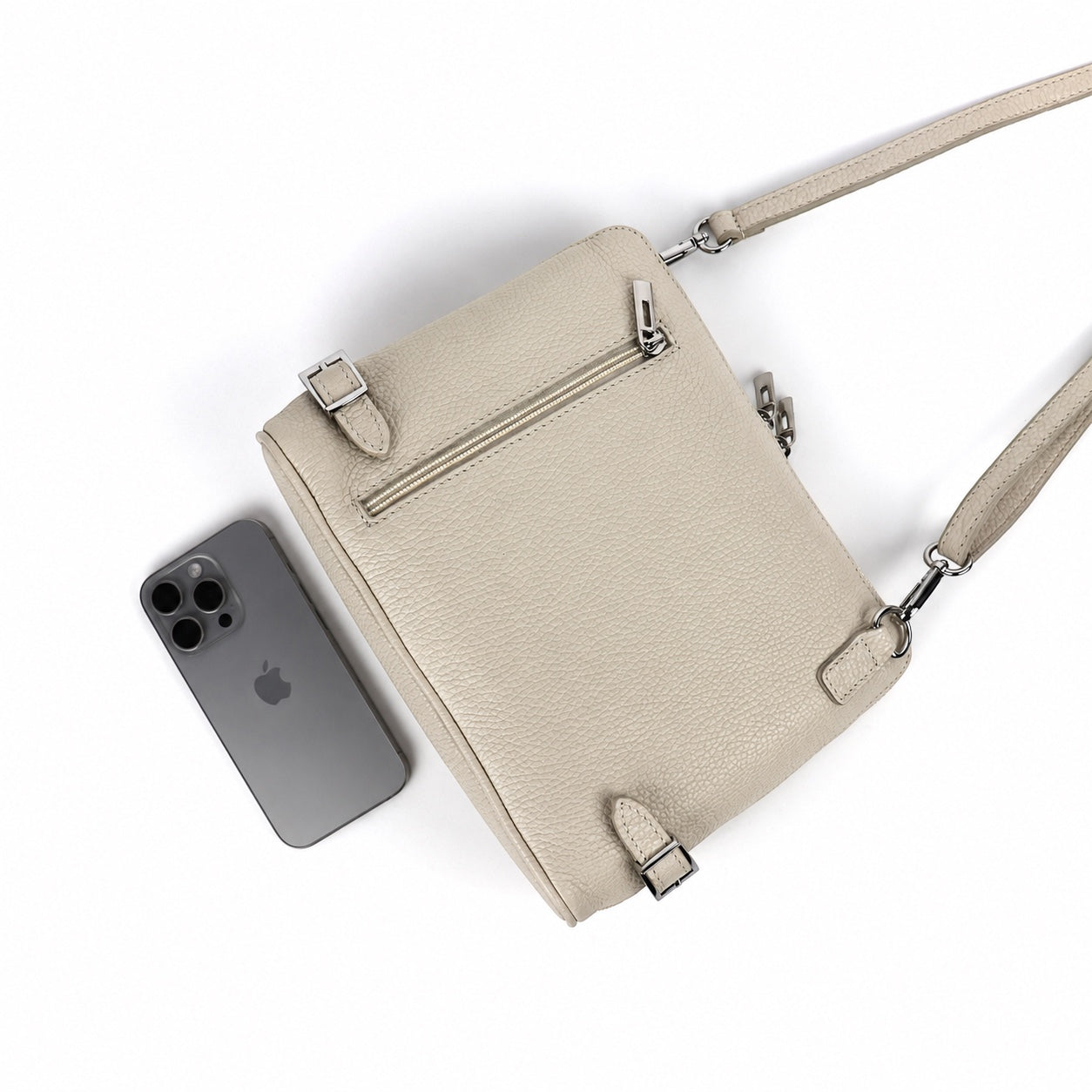 Eclectic Capri cream grained leather crossbody bag with rear zip pocket, side buckles, and smartphone for scale.