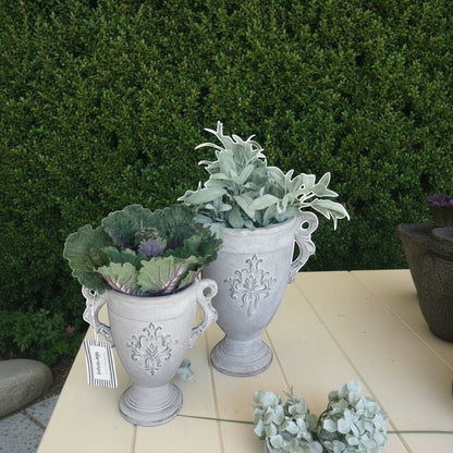 Eclectic antique cement trophy vase planters with ornate handles and weathered finish on a table with greenery