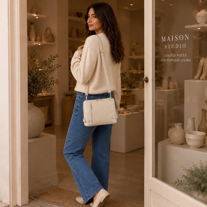 Woman wearing an Eclectic cream grained leather crossbody bag with side buckles and silver hardware in a boutique.