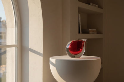 Eclectic crimson art glass vase with a sculptural curved silhouette and polished finish on a pedestal by a window