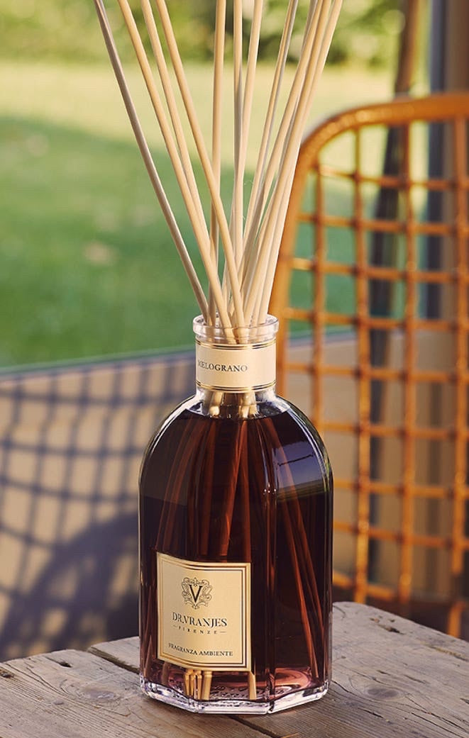 Eclectic Dr Vranjes Melograno reed diffuser in a glass bottle with natural sticks, set on a wooden table outdoors.