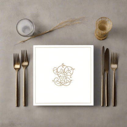 Eclectic Françoise Paviot white dinner napkin with gold monogram design, set on a table with gold cutlery.