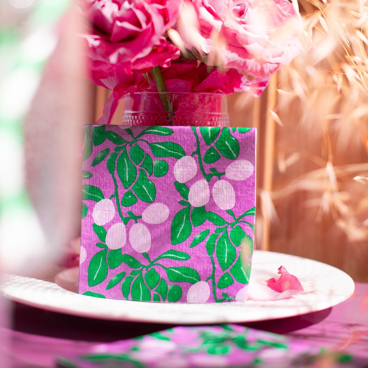 Eclectic Françoise Paviot Prune cocktail napkin with a purple plum tree print on a plate near pink roses
