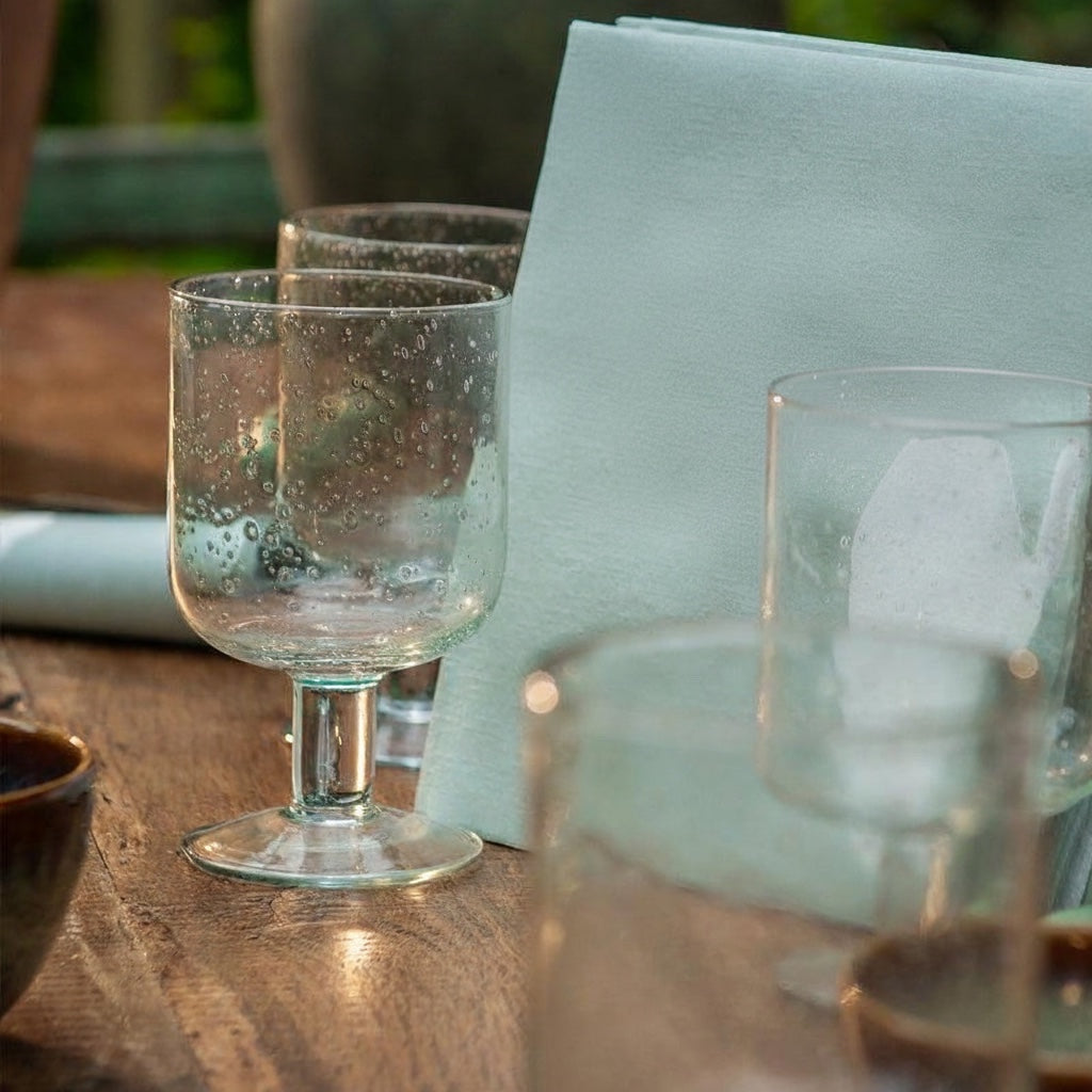 Eclectic Françoise Paviot sage green airlaid dinner napkin folded on a rustic wooden table with textured glass tumblers.