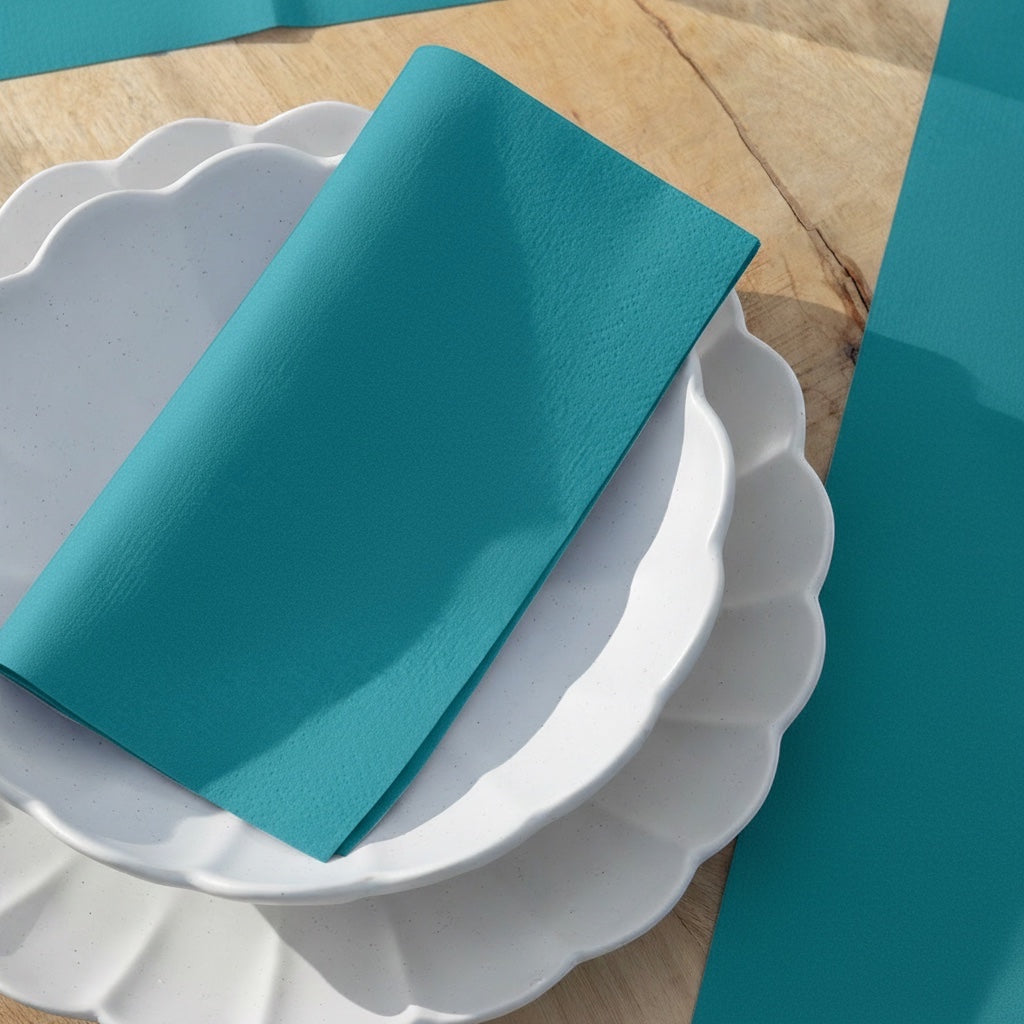 Eclectic Françoise Paviot deep teal airlaid dinner napkin folded on a white scalloped plate on a wooden table.