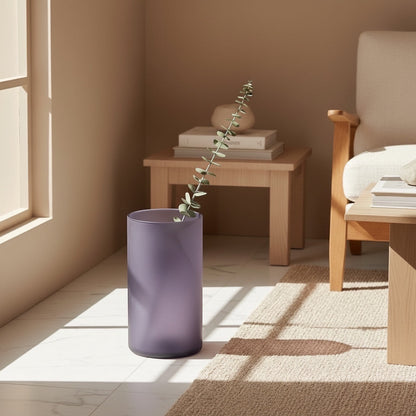Eclectic frosted plum glass cylinder vase standing on a floor with a single eucalyptus stem in a sunlit room.