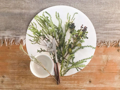 Eclectic round white placemat with green fynbos botanical print, styled on a wooden table with cutlery and a bowl.
