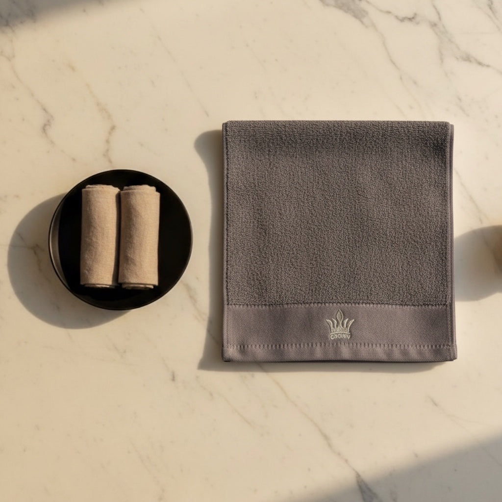 Eclectic grey cotton guest towel with embroidered crown detail on a marble surface next to rolled towels.