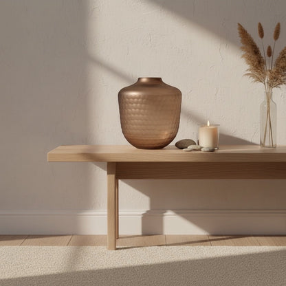 Eclectic hammered mauve glass vase on a wooden bench with a candle and dried grass in a sunlit room.
