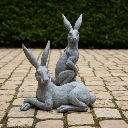 Eclectic Humphrey & Harriet cement hare garden sculptures in a weathered stone grey finish on a paved patio.