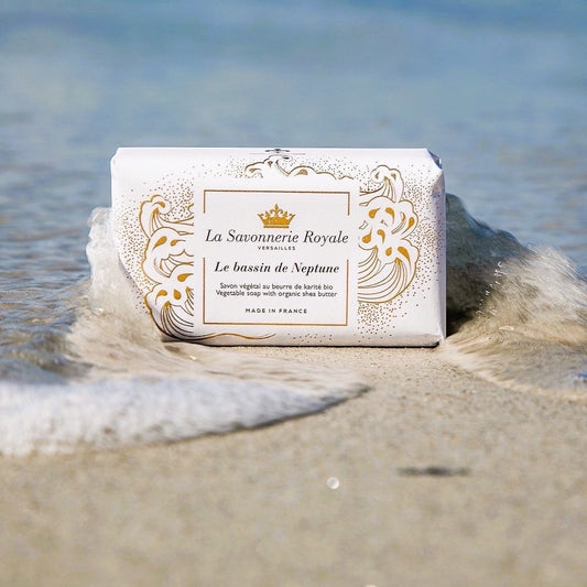 Eclectic La Savonnerie Royale Le Bassin de Neptune soap bar in decorative packaging resting on a sandy beach shore.