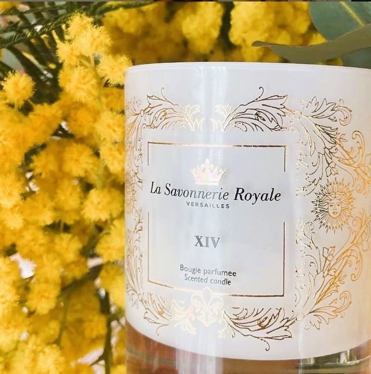 Eclectic La Savonnerie Royale XIV scented candle in a white glass vessel with ornate gold detailing near yellow flowers.