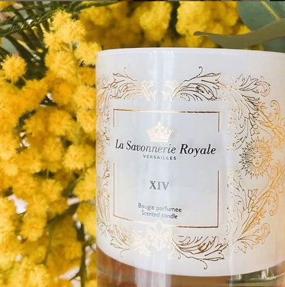 Eclectic La Savonnerie Royale XIV scented candle in a white glass vessel with ornate gold detailing near yellow flowers.