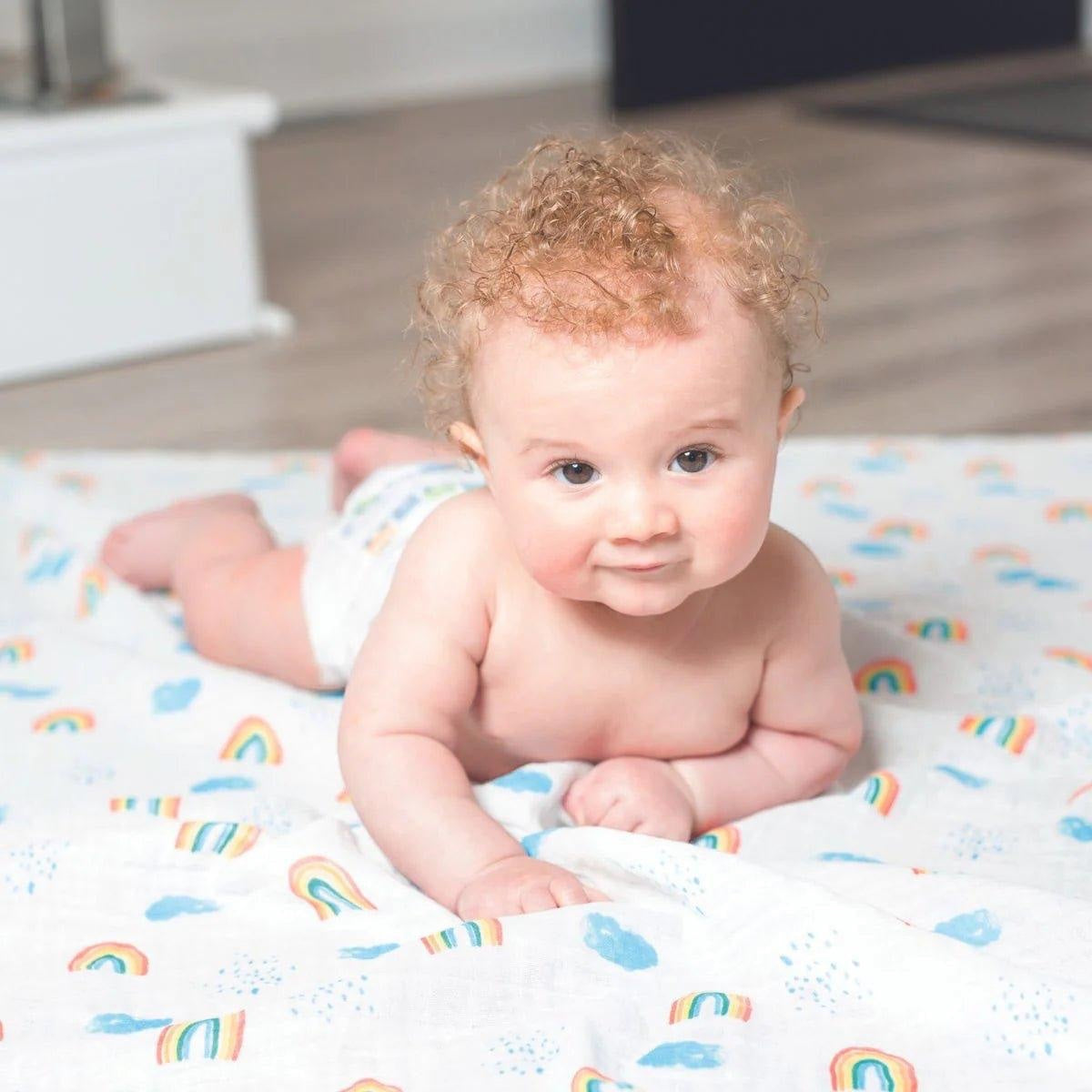 Eclectic Lulujo baby lying on a white cotton muslin swaddle with a colourful rainbow and sky print