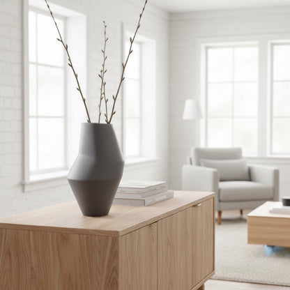 Eclectic tall matte grey faceted ceramic vase holding thin branches on a light wood sideboard in a modern living room