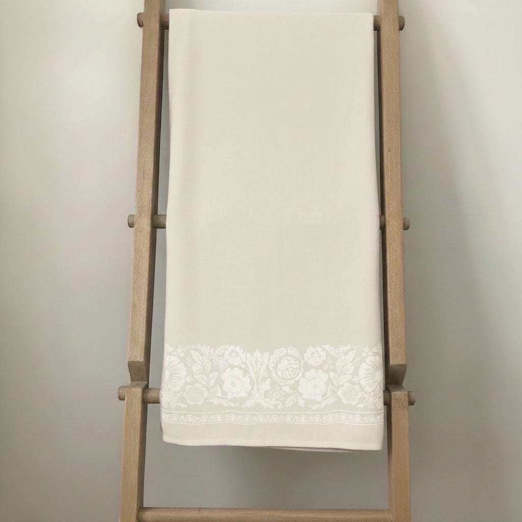 Eclectic natural beige linen-look tablecloth featuring an intricate white floral border draped on a wooden ladder.