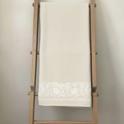 Eclectic natural beige linen-look tablecloth featuring an intricate white floral border draped on a wooden ladder.