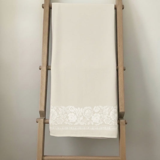 Eclectic natural beige linen-look tablecloth featuring an intricate white floral border draped on a wooden ladder.