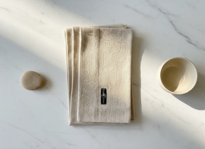 Eclectic set of four natural stonewashed linen napkins with a tonal scroll pattern on a white marble surface.