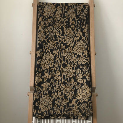 Eclectic Noir Baroque round tablecloth in black and gold with an intricate floral pattern draped over a wooden rack.