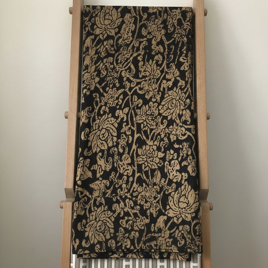 Eclectic Noir Baroque round tablecloth in black and gold with an intricate floral pattern draped over a wooden rack.