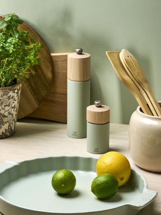 Eclectic Peugeot Boreal sage green beechwood salt and pepper mills on a kitchen counter with fresh fruit and utensils.