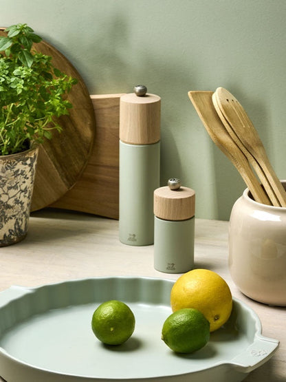 Eclectic Peugeot Boreal sage green beechwood salt and pepper mills on a kitchen counter with fresh fruit and utensils.