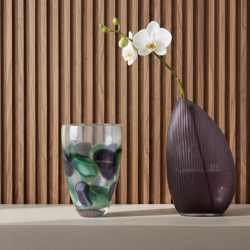 Eclectic purple and green petal glass vases on a shelf against a wooden slatted wall with a white orchid stem.