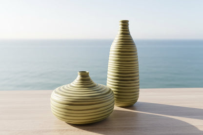 Eclectic tall ribbed olive glass vase and matching wide vase on a wooden surface against an ocean backdrop.