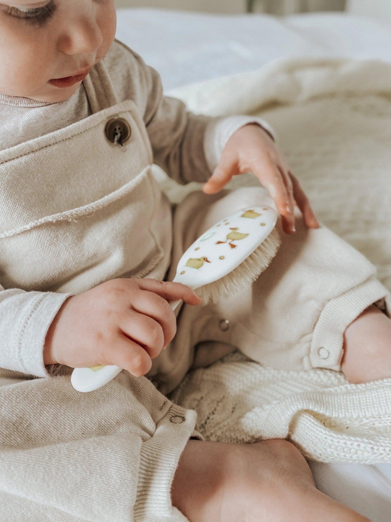 Eclectic Rock & Ruddle baby hairbrush with duckling design held by a baby in beige overalls on a soft blanket