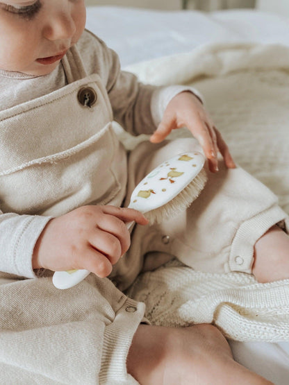 Eclectic Rock & Ruddle baby hairbrush with duckling design held by a baby in beige overalls on a soft blanket