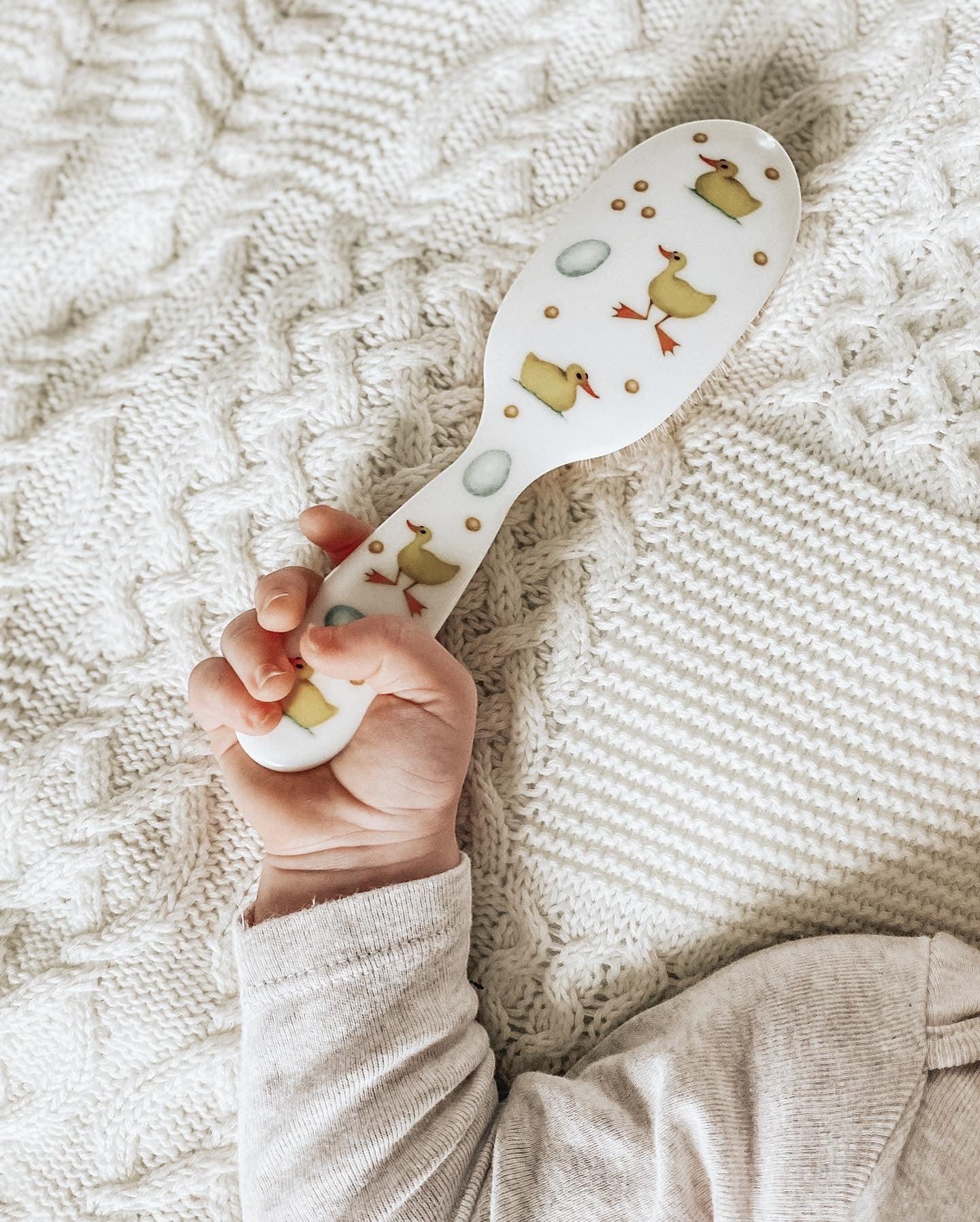 Eclectic Rock & Ruddle baby hairbrush with duckling and blue egg pattern held in a baby's hand on a white blanket.