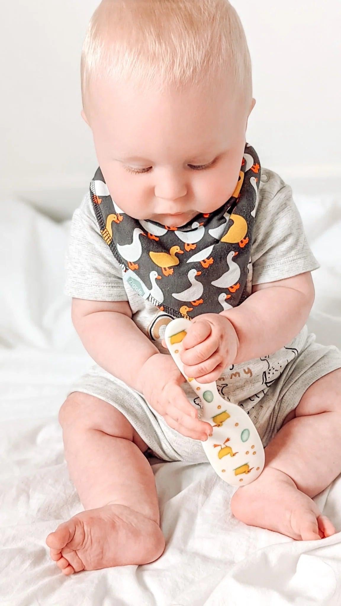Eclectic Rock & Ruddle baby hairbrush with duckling design held by a baby wearing a patterned bib on a white bed