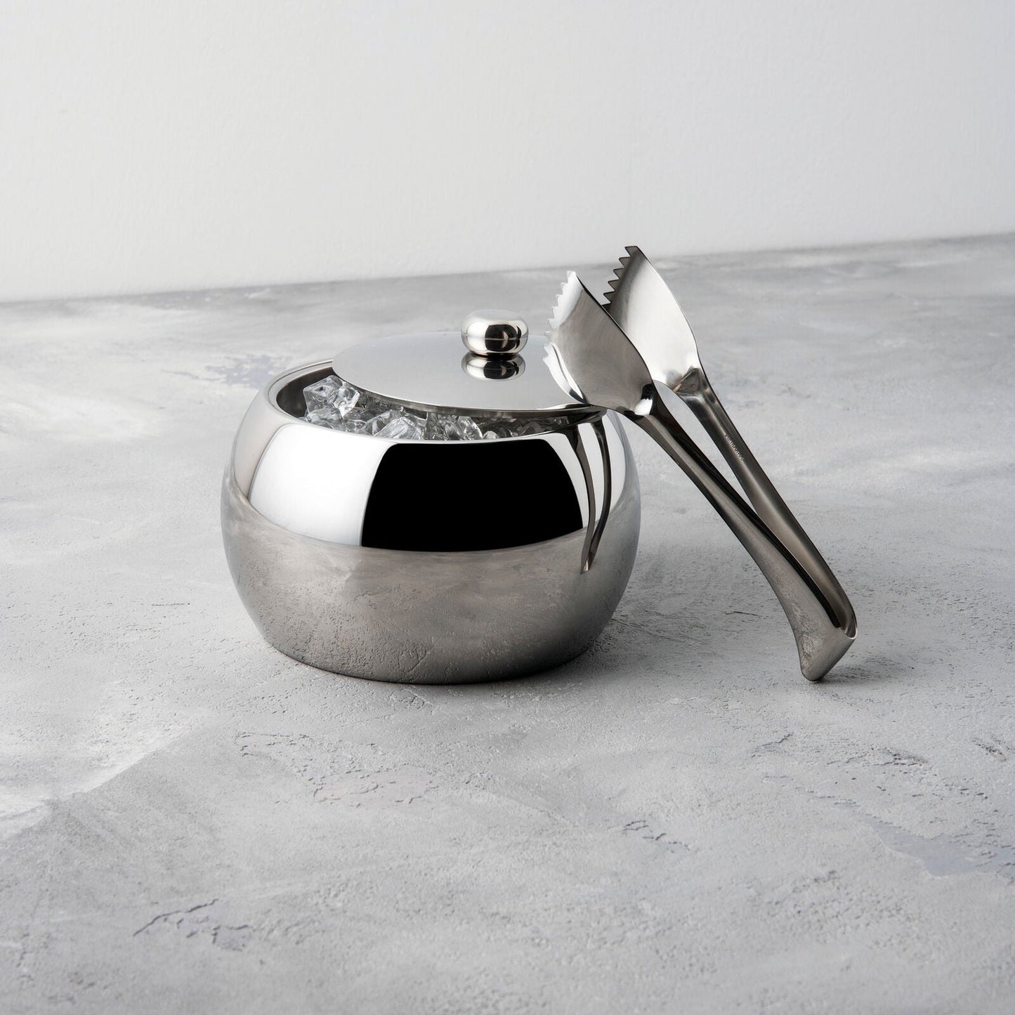 Eclectic Sambonet Living stainless steel ice bucket and tongs with a mirror-polished finish on a grey stone surface.