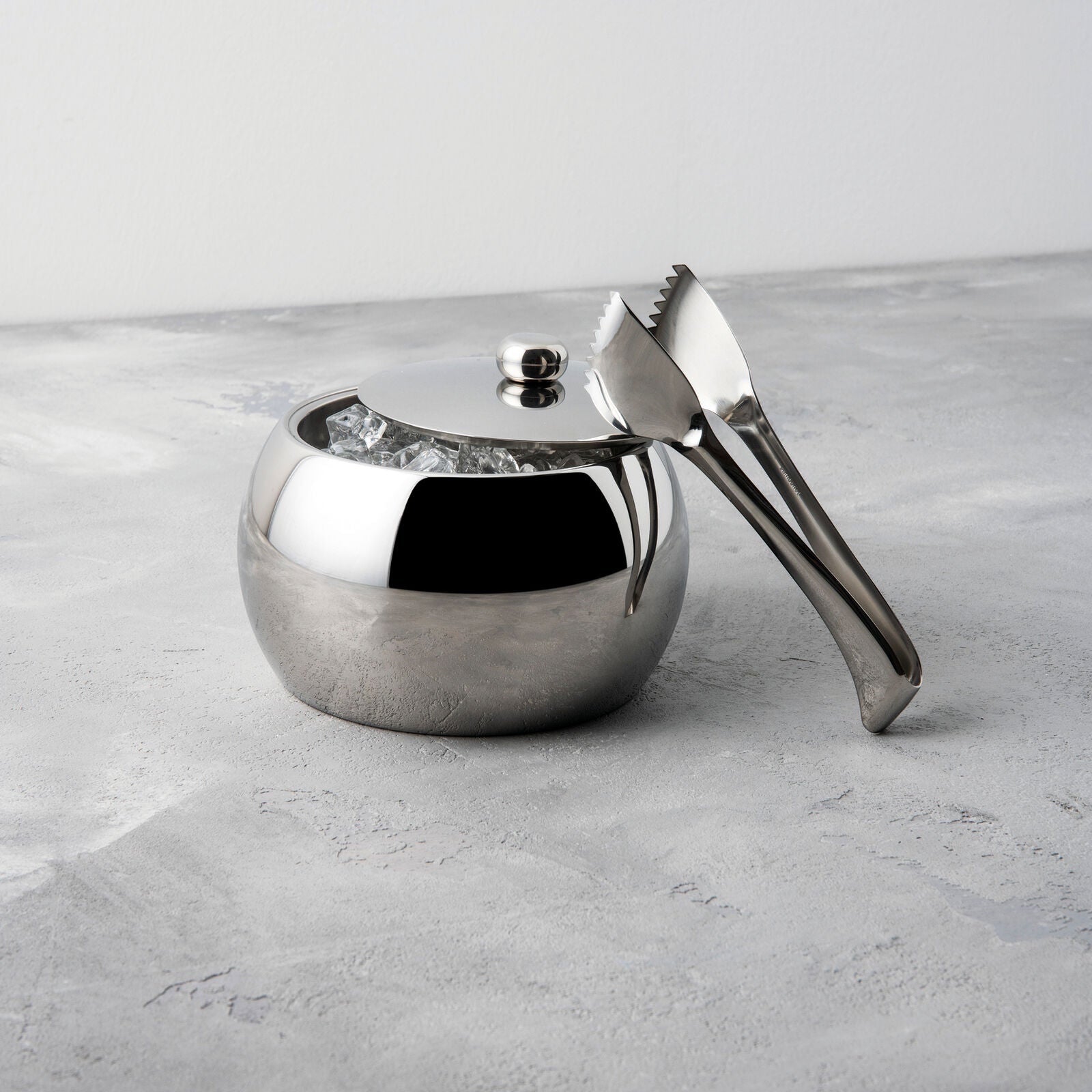 Eclectic Sambonet Living stainless steel ice bucket and tongs with a mirror-polished finish on a grey stone surface.