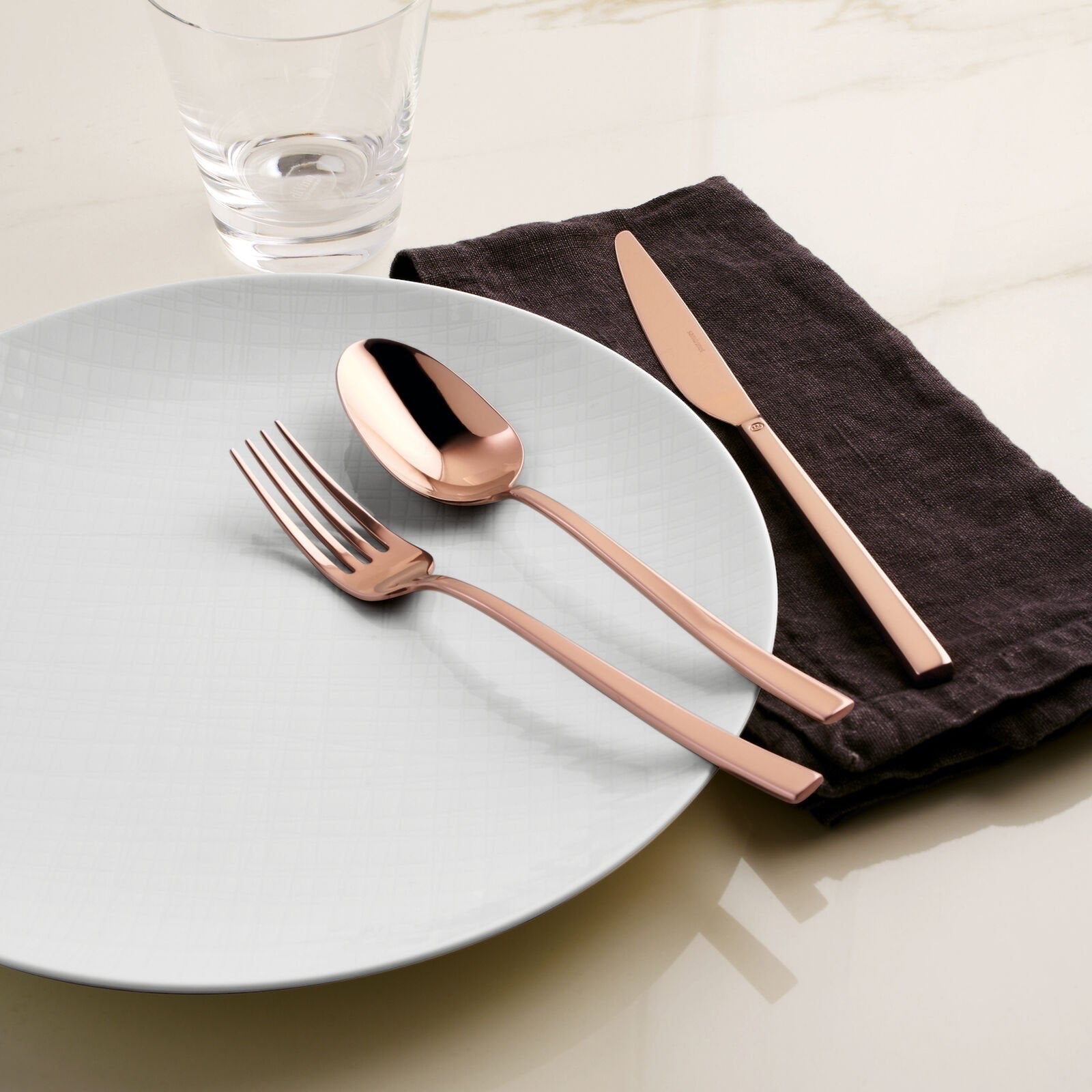 Eclectic Sambonet Rock mirror copper cutlery set with fork, spoon, and knife on a white plate and dark linen napkin.