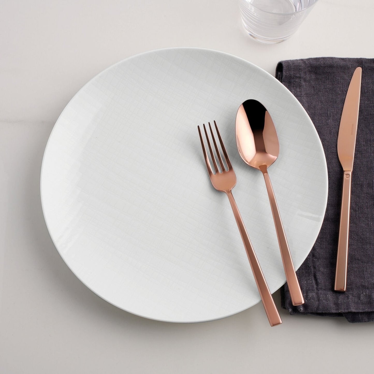 Eclectic Sambonet Rock mirror copper cutlery set with fork, spoon, and knife on a white plate and dark grey napkin.
