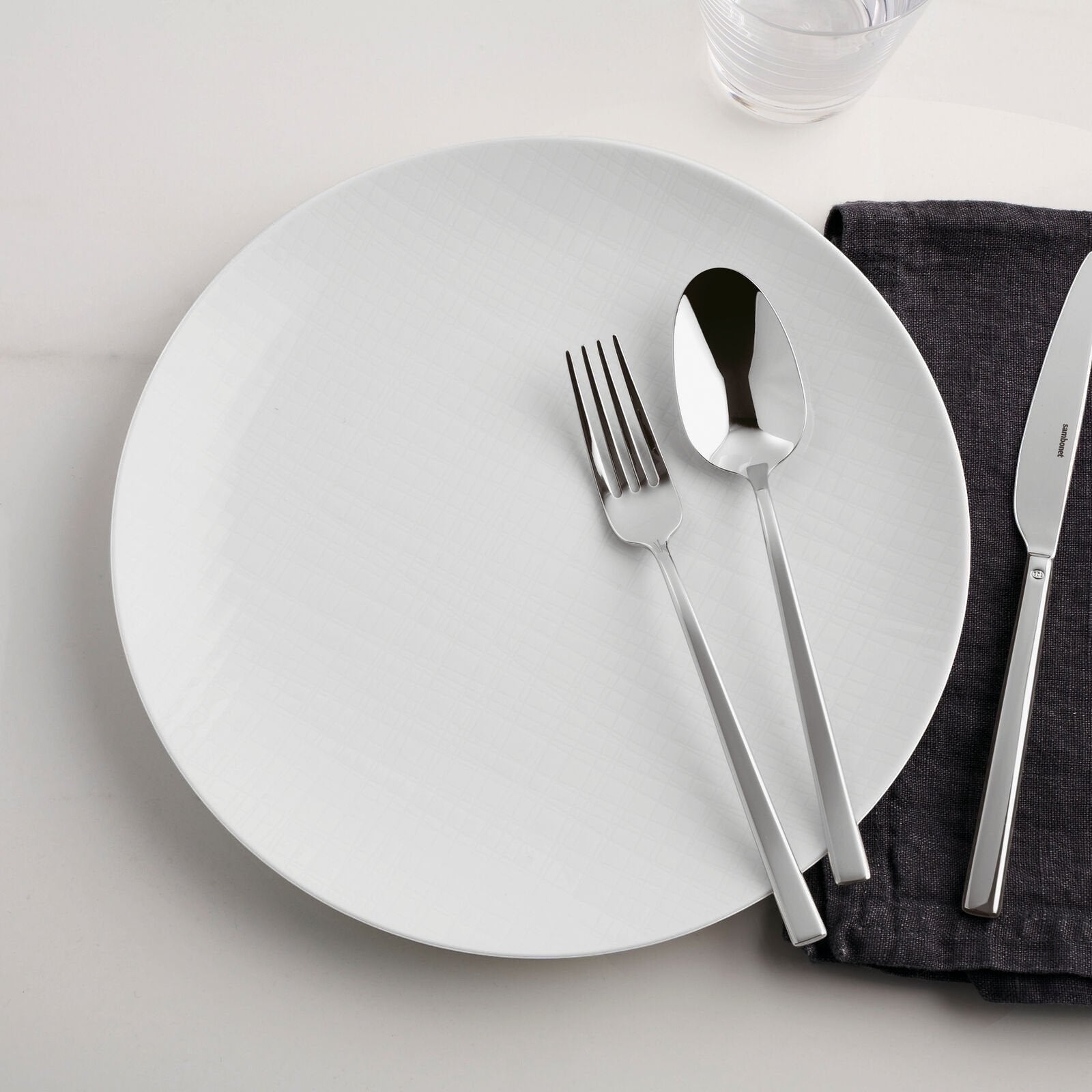 Eclectic Sambonet Rock Mirror stainless steel fork, spoon, and knife set on a white plate with a dark grey napkin.