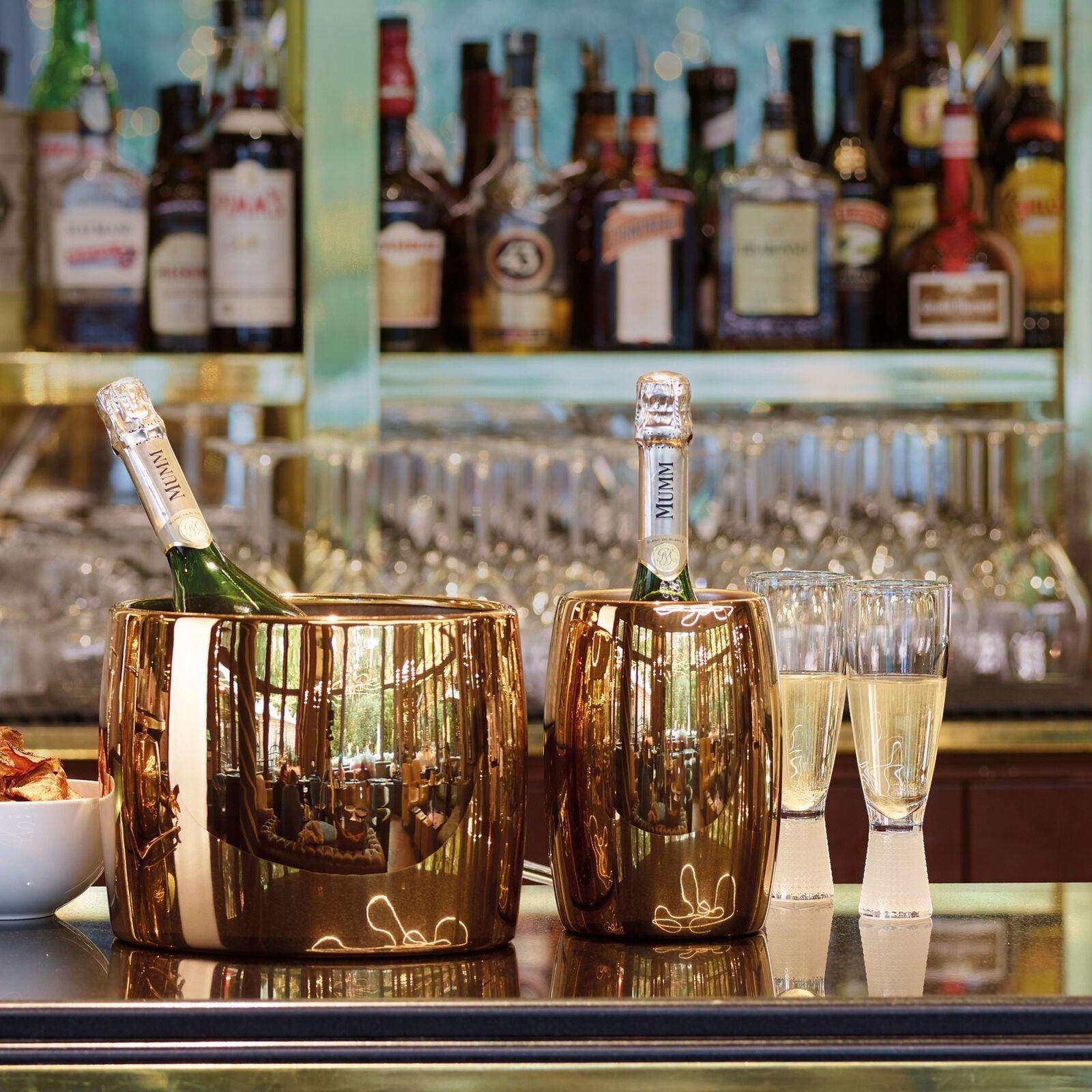 Eclectic Sambonet Sphera oval wine cooler in PVD cognac stainless steel holding champagne bottles on a bar counter.