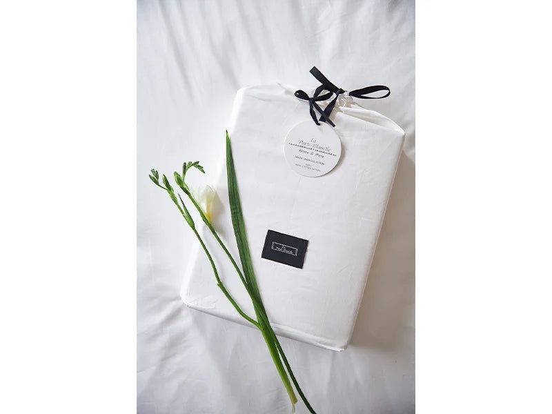Eclectic Saxon white cotton sateen duvet set in a branded drawstring storage bag with a fresh white flower stem.