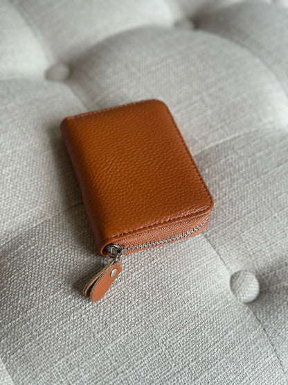 Eclectic tan pebbled leather zip-around card holder with silver hardware resting on a tufted fabric surface.
