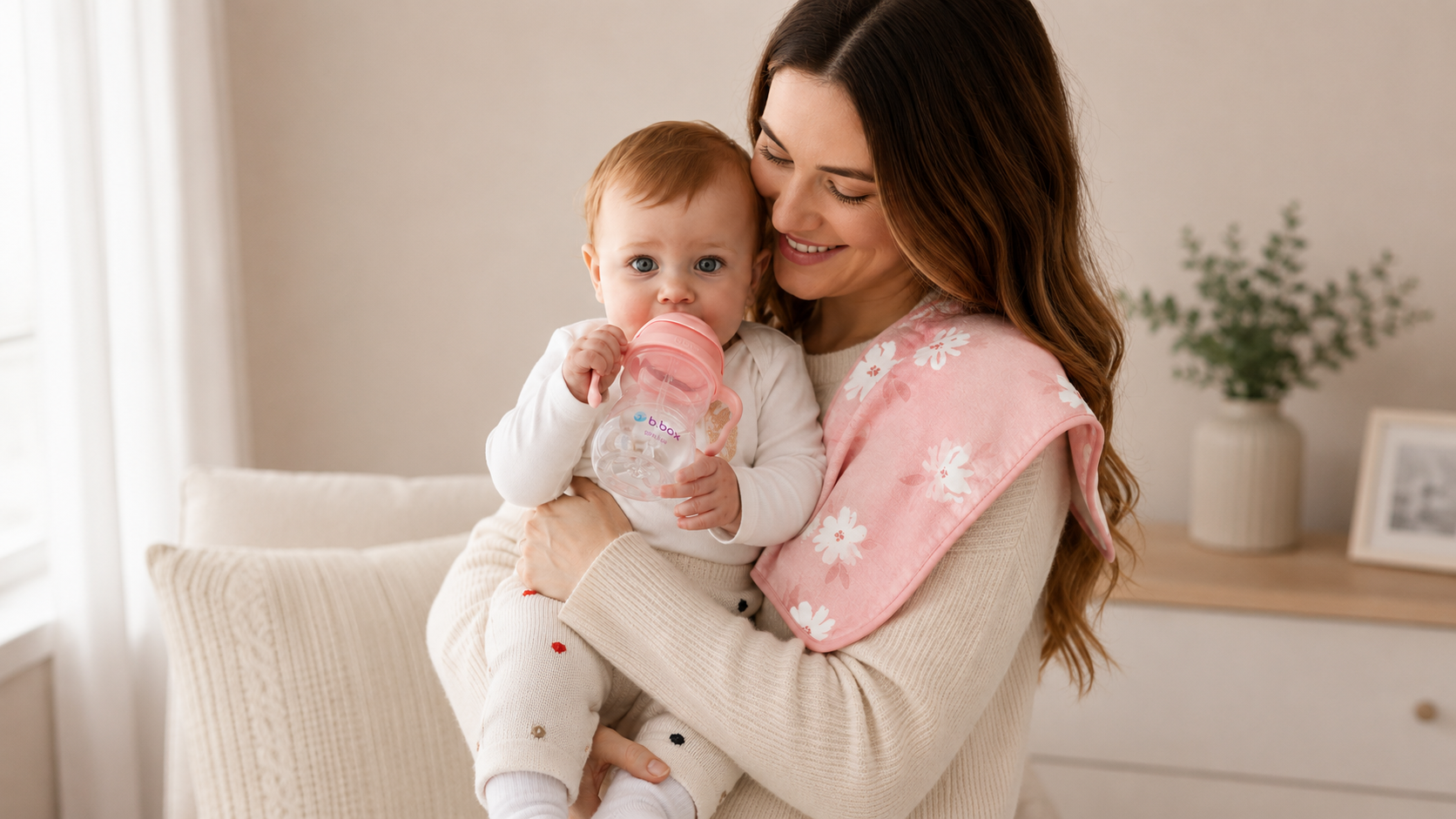 Baby drinking from a sippy cup while being held by a parent wearing a burp cloth.