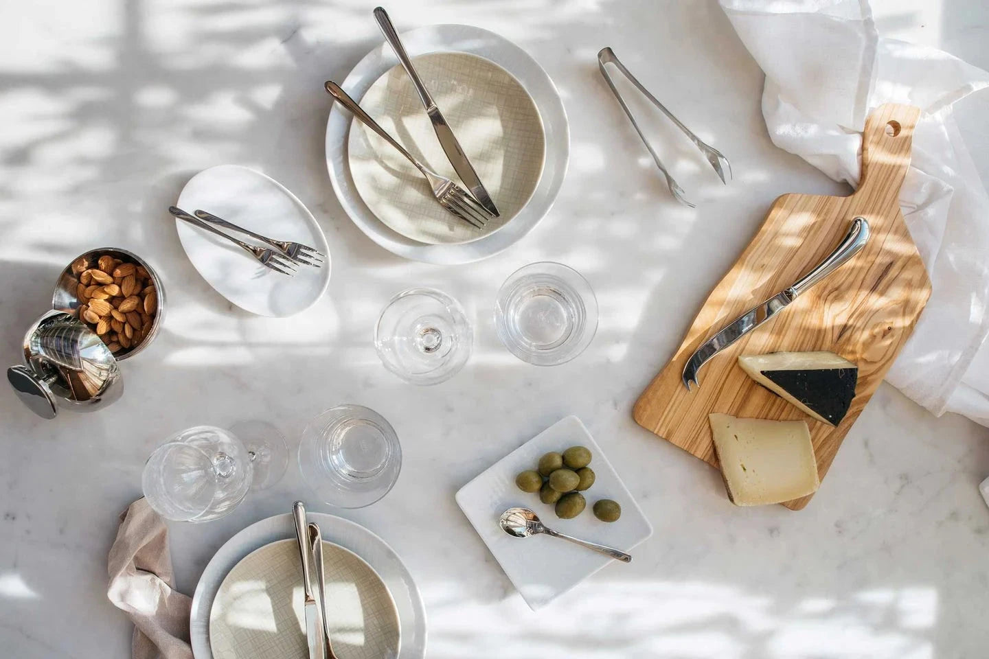 Flatware and serving utensils arranged on a marble table with plates, glasses, and cheese.