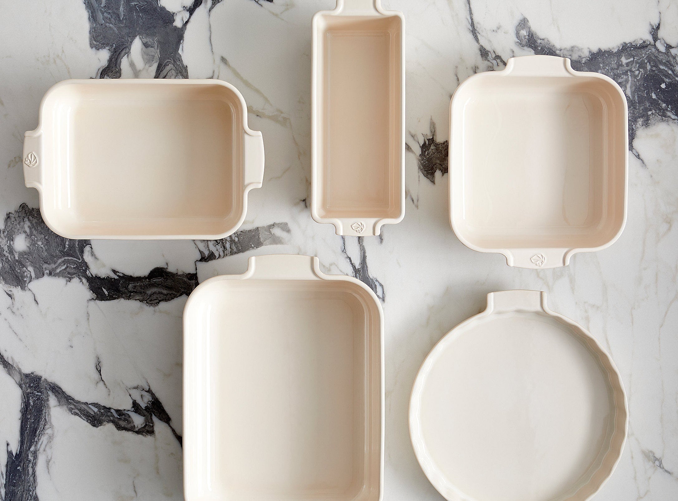 Cream ovenware dishes and baking trays arranged on a marble surface.
