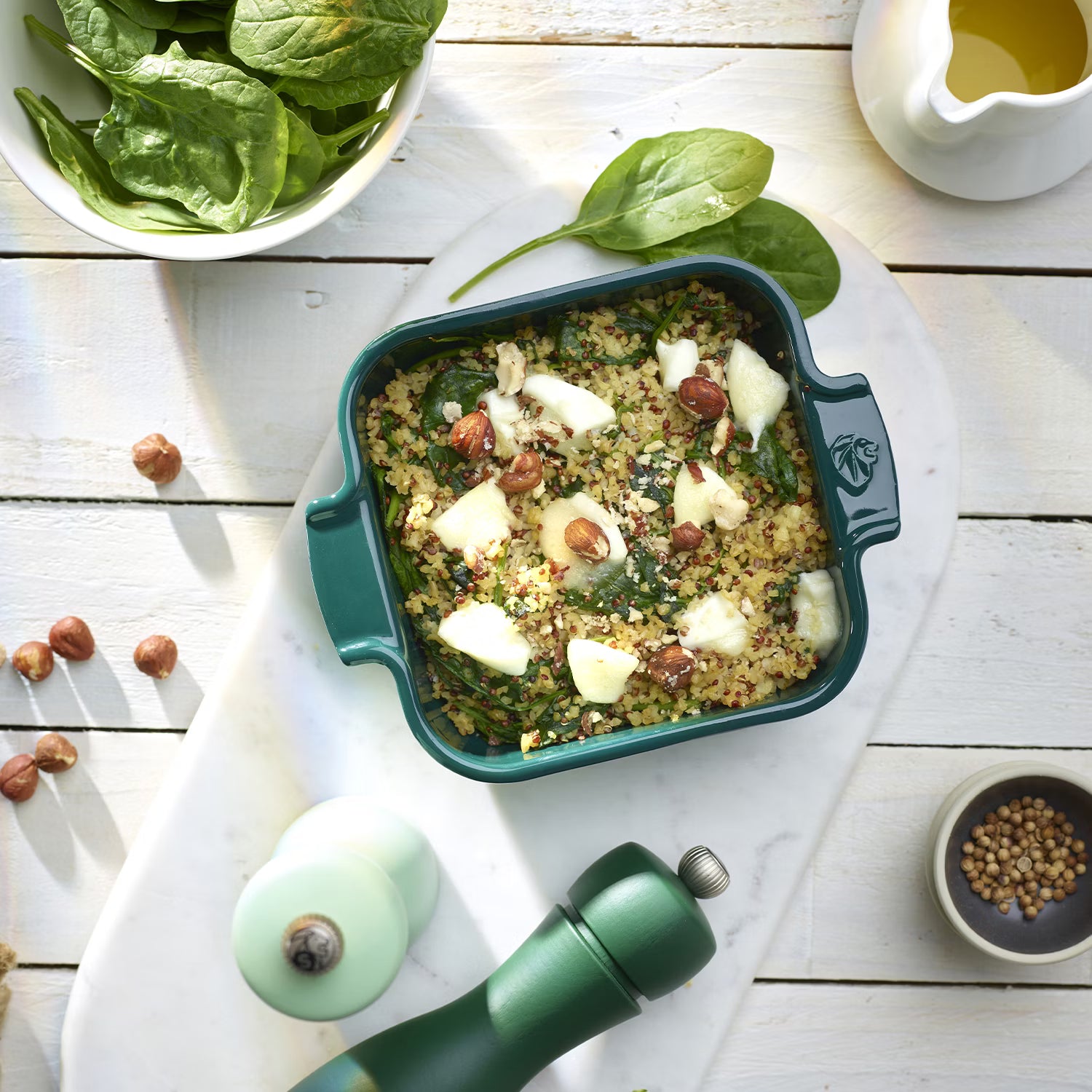 Green roasting and baking dish filled with grains, spinach, nuts, and cheese on a white table.