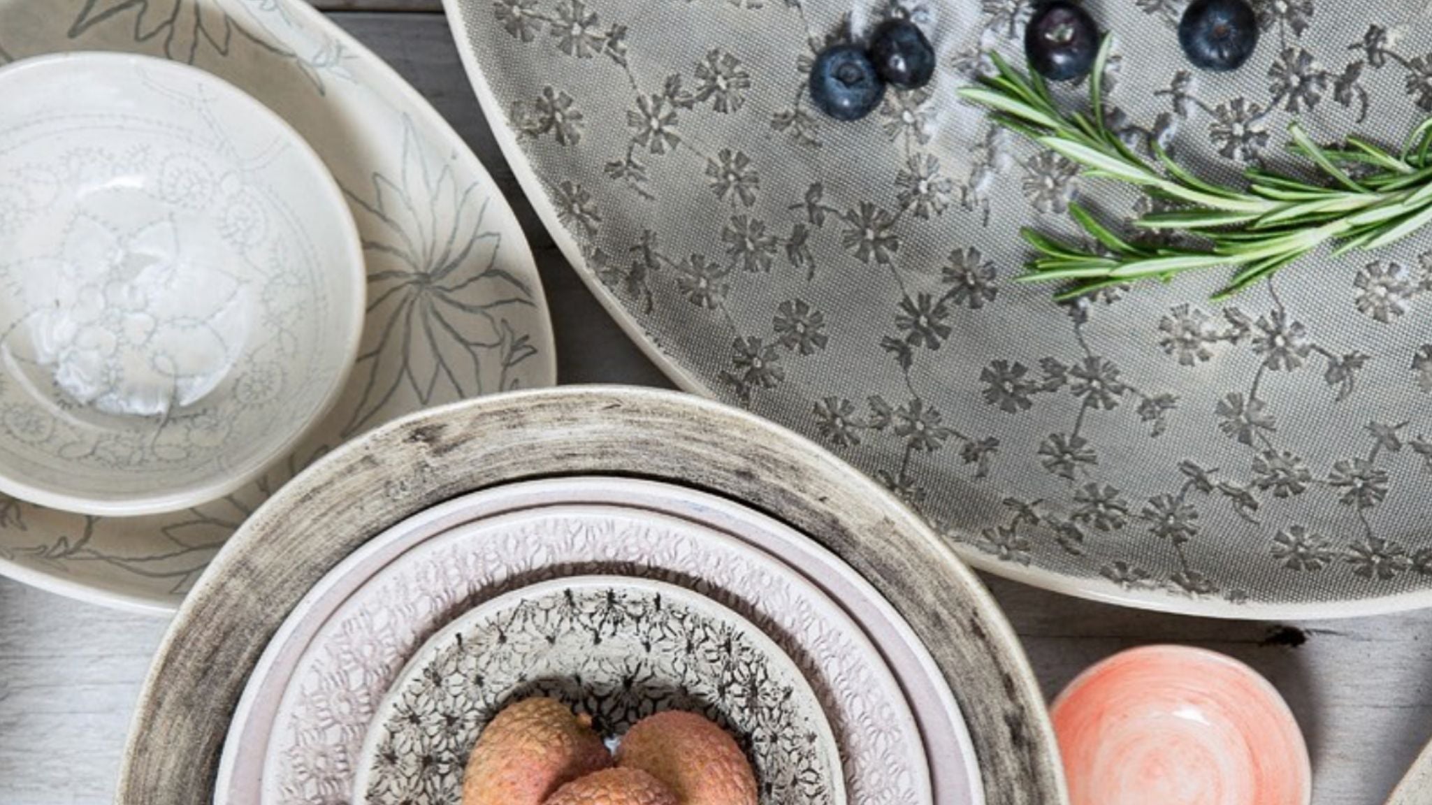 Serveware collection with patterned bowls, trays, serving dishes, and rosemary on a styled tabletop.