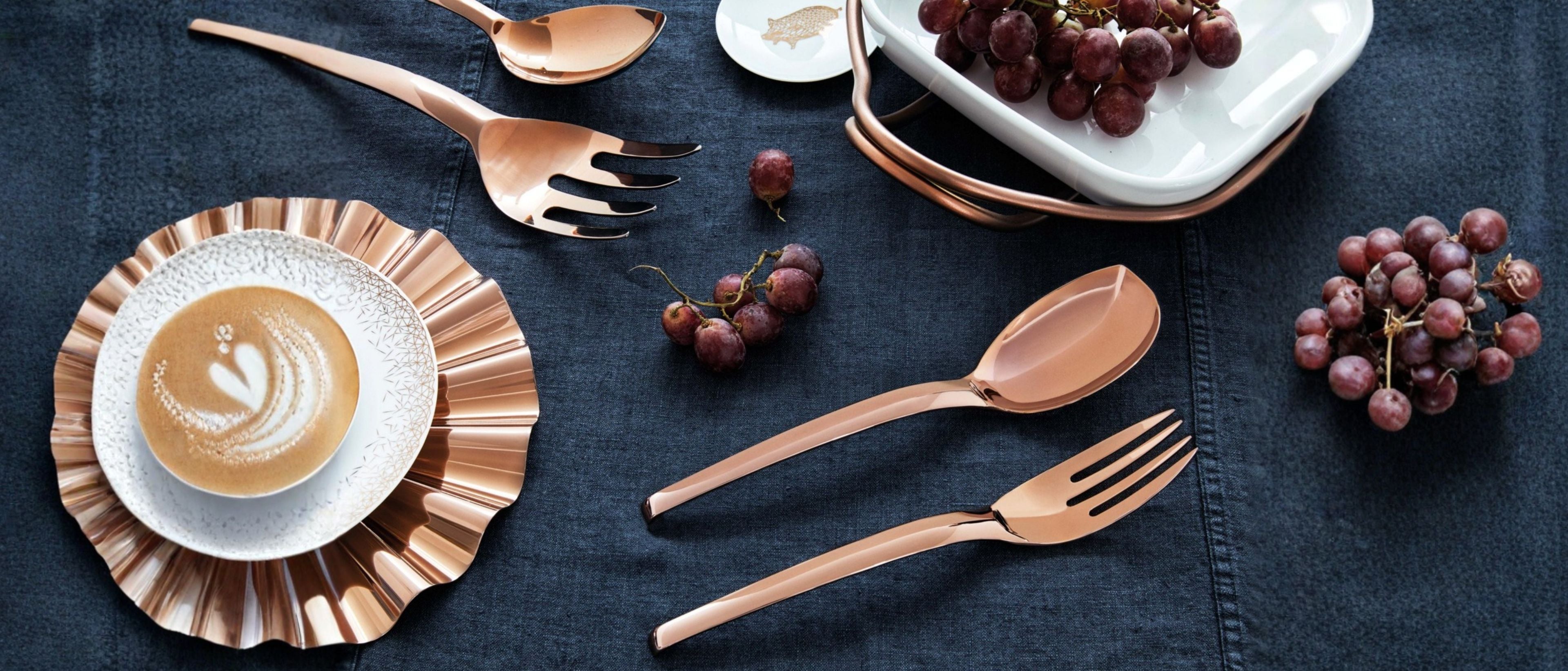 Rose gold serving utensils arranged with grapes, tableware, and a coffee cup on a dark linen table setting.
