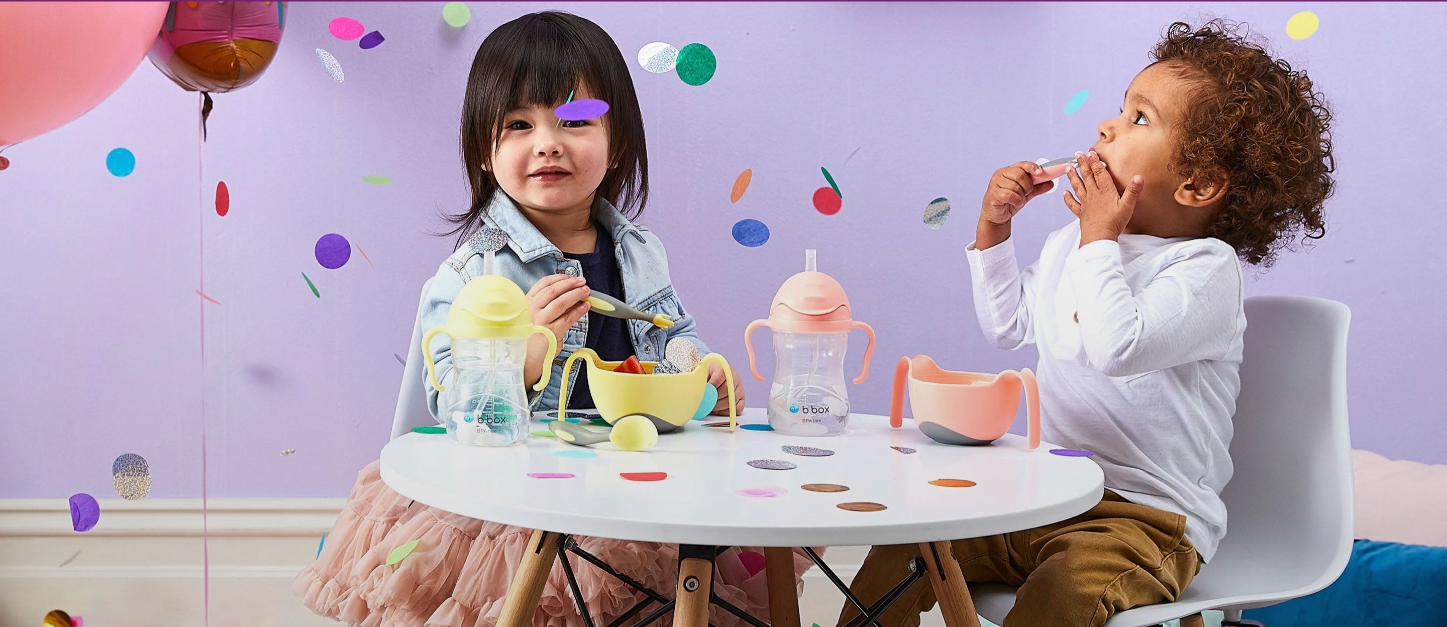 Two children using sippy cups at a table with colourful party decorations.