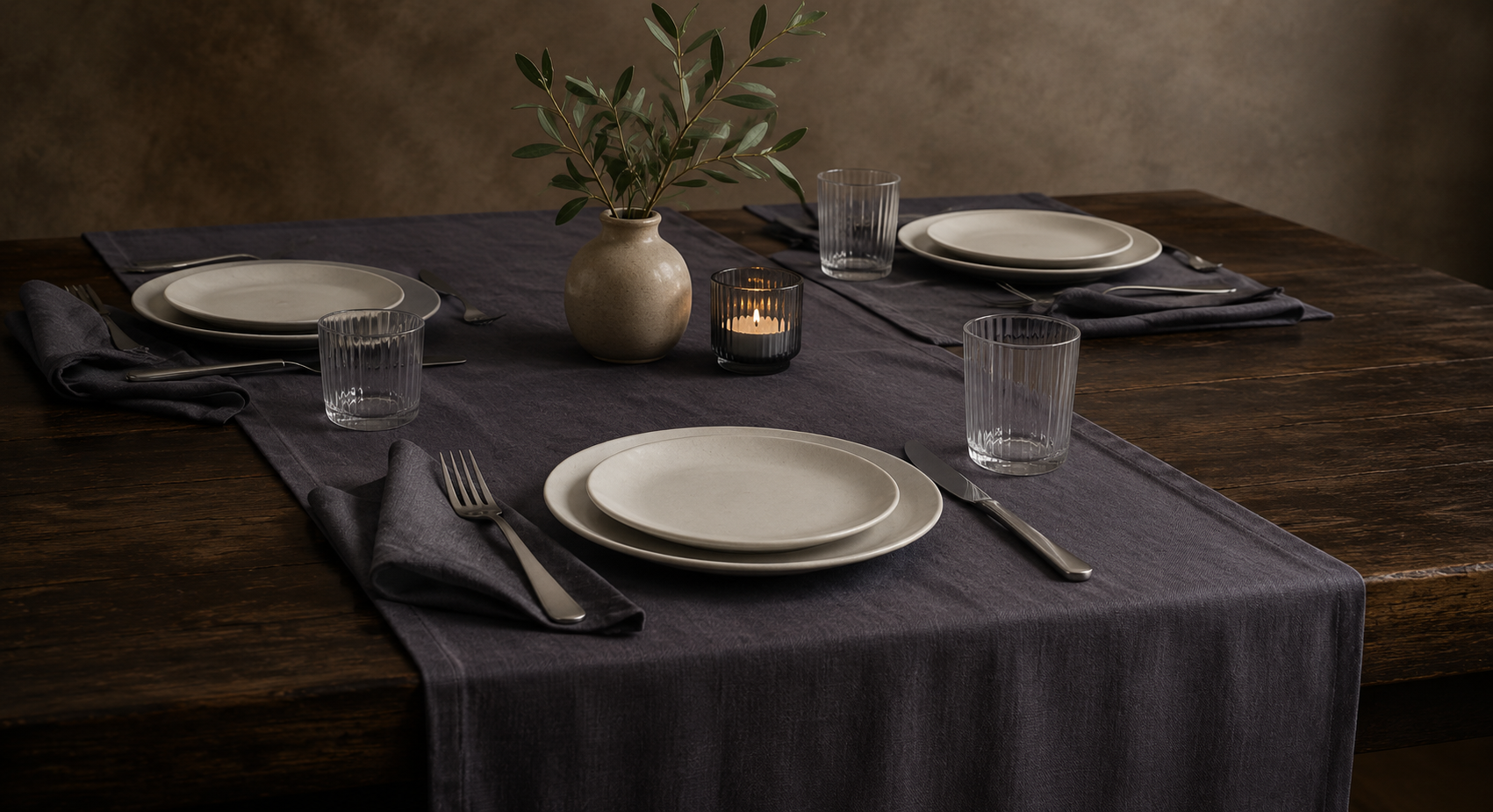 Dark table runner styled with dinner plates, napkins, glassware, cutlery, a candle, and a vase on a wooden table.